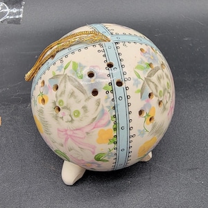 Puede incluir: Una esfera de cerámica blanca con un diseño estampado en azul y negro y una cuerda dorada. La esfera tiene agujeros y está decorada con un patrón floral y una cara de gatito. La esfera está sentada sobre tres pequeños pies.