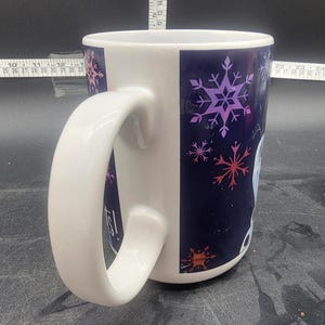Puede incluir: Taza de cer&aacute;mica blanca con un dise&ntilde;o de copo de nieve azul y morado. La taza tiene una asa grande y dice "Let It Go!" en el lateral.