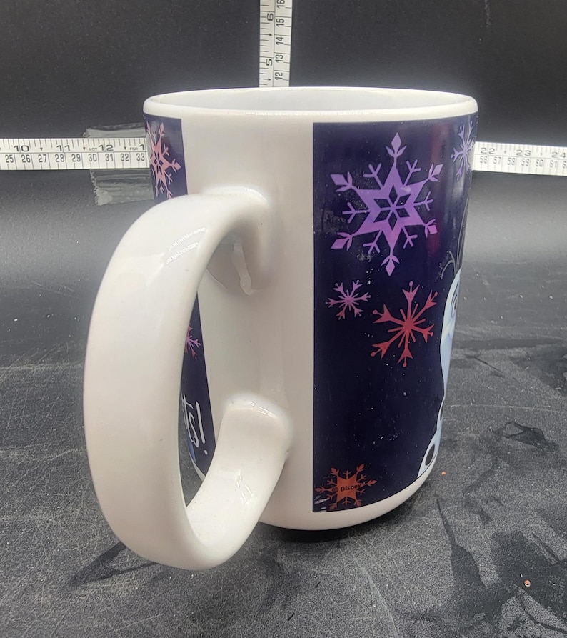 Puede incluir: Taza de cer&aacute;mica blanca con un dise&ntilde;o de copo de nieve azul y morado. La taza tiene una asa grande y dice "Let It Go!" en el lateral.