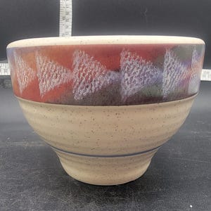 May include: A beige ceramic bowl with a multi-colored geometric design around the rim. The design features shades of red, orange, purple, and white.