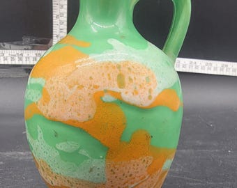 May include: A green ceramic pitcher with a handle and a spout. The pitcher is decorated with orange and white abstract patterns. The base is a solid green color.