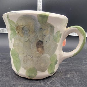 Puede incluir: Una taza de cerámica blanca con un diseño floral con hojas verdes y flores grises. La taza tiene un asa y un borde rosa claro.