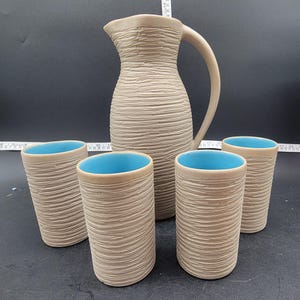 May include: A set of five ceramic pieces in a light brown colour with a textured surface. The set includes a jug and four cups. The cups have a light blue interior.
