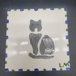 May include: A hand-painted ceramic tile with a gray cat silhouette and green eyes. The tile has a white background and a blue and green border with dots.