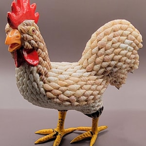 Puede incluir: Una figura de gallo blanco hecha completamente de conchas marinas. El gallo tiene una cresta y una papada rojas, patas amarillas y una cola marrón.