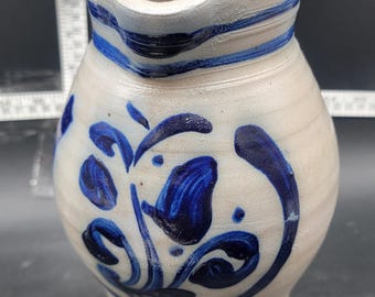 May include: A white ceramic pitcher with blue floral designs and blue stripes around the rim. The pitcher has a rounded body and a slightly flared spout.