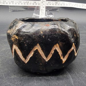 Puede incluir: Una pequeña maceta de cerámica negra con una superficie rugosa y texturizada. La maceta tiene un esmalte blanco y un patrón geométrico de zigzags alrededor del borde.