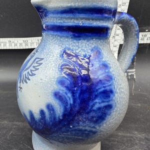 May include: A blue and white ceramic pitcher with a handle. The pitcher features a dark blue design on a light blue background. The design includes a bird and abstract patterns. The pitcher is approximately 8 inches tall.