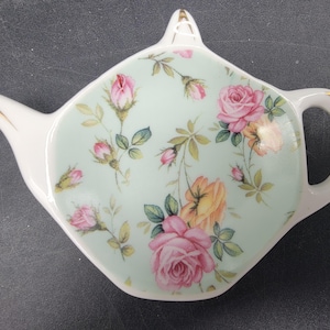May include: A white ceramic teapot-shaped tea bag holder with a light blue background and a floral pattern of pink and yellow roses.