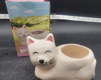 May include: White ceramic cat planter with a smiling face and pink ears. The planter has a small hole in the top for drainage. The planter is sitting on a table with a ruler in the background. The box says "Plants not included, plantas nun incluian".