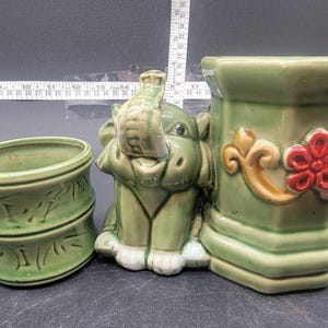 May include: A set of three green ceramic planters featuring a whimsical elephant design. The planters are decorated with floral motifs and have a textured finish.