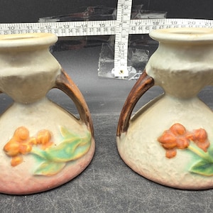Puede incluir: Dos jarrones de cerámica con base beige, asas marrones y detalles florales naranjas y verdes. Los jarrones tienen una forma única y probablemente son artículos decorativos.