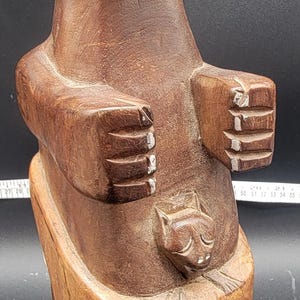 Op de afbeelding: Een houten sculptuur van een kat die een kleinere kat in zijn armen houdt. Het beeld is bruin en heeft een natuurlijke houten afwerking. De ogen van de kat zijn wit geschilderd.