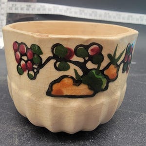 Puede incluir: Una pequeña maceta de cerámica blanquecina con base acanalada y diseño floral. El diseño presenta racimos de bayas rojas y moradas, hojas verdes y detalles en naranja. La maceta tiene un aspecto ligeramente agrietado y envejecido.