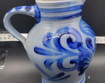 May include: A blue and white ceramic pitcher with a blue floral design. The pitcher has a handle and a spout.