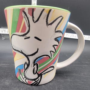 May include: White ceramic mug with a colorful striped design and a black and white illustration of Snoopy. The mug has a green rim and a white handle.