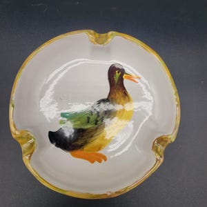 以下が含まれることがあります： 黄色い縁取りのある白い陶器製の灰皿で、アヒルの絵が描かれています。アヒルは黄色、緑、黒で、オレンジ色のくちばしをしています。