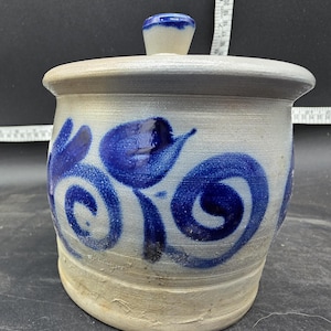 Puede incluir: Un tarro de cerámica hecho a mano con tapa. El tarro es de color beige claro con diseños florales azules y un pomo azul en la tapa. El tarro tiene un aspecto rústico y artesanal, adecuado para la decoración de la cocina o del hogar.