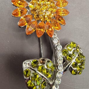 May include: A rhinestone sunflower brooch with orange and yellow petals, green leaves, and clear rhinestones.