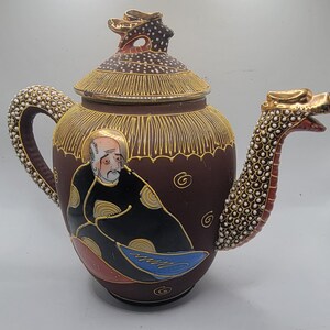 Puede incluir: Una tetera de cerámica marrón con un pico de dragón y una tapa con un diseño dorado y blanco. La tetera presenta una imagen pintada de un hombre con una túnica negra.