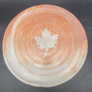 May include: A white ceramic plate with a brown glaze and a white leaf design in the center. The plate has a concentric circle pattern.