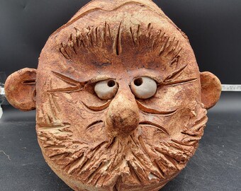 May include: A ceramic sculpture of a face with a large nose, exaggerated eyebrows, and a bushy beard. The sculpture is painted in shades of brown and has a textured surface.
