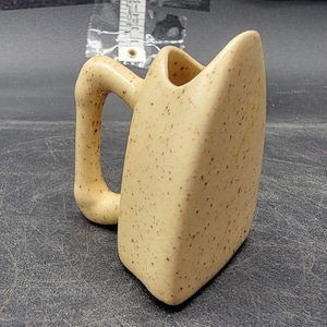 Puede incluir: Una taza de cerámica beige con un diseño angular único. La taza tiene un cuerpo rectangular con un asa curva y está moteada con manchas marrones. La parte superior de la taza tiene una forma puntiaguda distintiva.