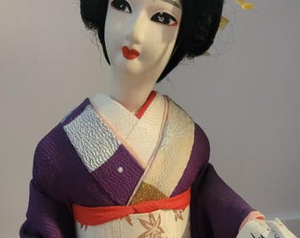 Pode incluir: Uma boneca japonesa tradicional vestindo um quimono roxo e branco com uma faixa vermelha. A boneca tem cabelo preto e um rosto branco com lábios vermelhos. A boneca está segurando um pergaminho com escrita japonesa.