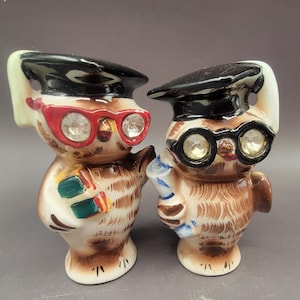 May include: A pair of ceramic owl salt and pepper shakers. The owls are wearing graduation caps and glasses with rhinestones. One owl is holding a book and the other is holding a diploma.