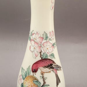 May include: A white ceramic vase with a gold rim and a floral design featuring pink flowers, green leaves, and a red bird perched on a branch with a pomegranate.