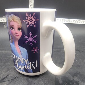 Puede incluir: Taza de cer&aacute;mica blanca con un dise&ntilde;o azul y morado que presenta una imagen de dibujos animados de Elsa de Frozen. La taza tiene un asa y el texto "Destiny Awaits!" impreso en la parte delantera.