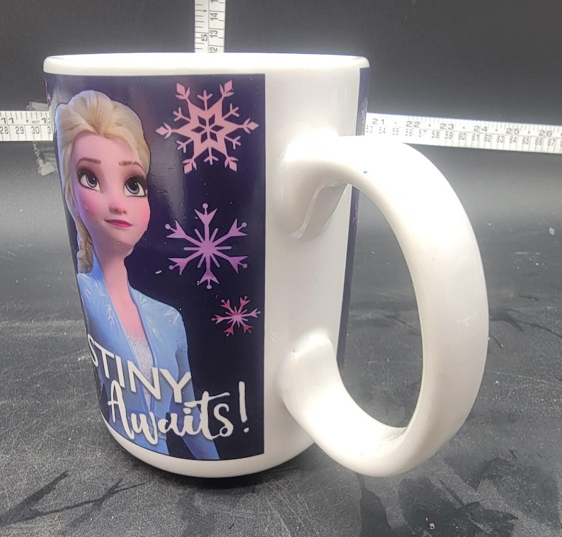 Puede incluir: Taza de cer&aacute;mica blanca con un dise&ntilde;o azul y morado que presenta una imagen de dibujos animados de Elsa de Frozen. La taza tiene un asa y el texto "Destiny Awaits!" impreso en la parte delantera.