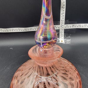 Può includere: Una bottiglia di vetro rosa con un design a spirale bianco e un tappo di vetro iridescente. La bottiglia è alta circa 15 cm.