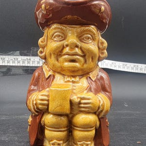 May include: A ceramic Toby jug figurine depicting a seated man in a brown hat and coat, holding a mug. The jug is primarily yellow and brown, with intricate details and a glossy finish. A vintage collectible.