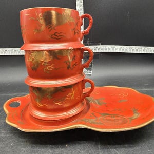May include: A stack of three red teacups with gold dragon designs and matching saucer. The cups and saucer have a gold trim. The teacups are stacked on top of each other. The saucer has a unique shape.
