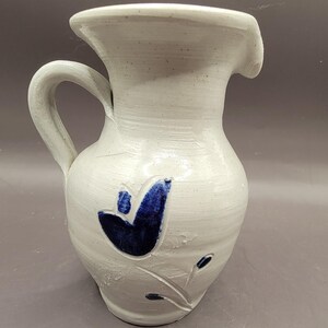 May include: A white ceramic pitcher with a blue floral design. The pitcher has a handle and a spout.