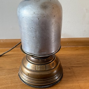 Könnte beinhalten: Vintage Haartrockner mit einer Kuppel aus gebürstetem Metall und einem Messingfuß. Die Kuppel hat kleine Belüftungslöcher. Der Sockel hat ein geriffeltes Design und ein kleines rechteckiges Schild. Ein Netzkabel geht vom Sockel aus.