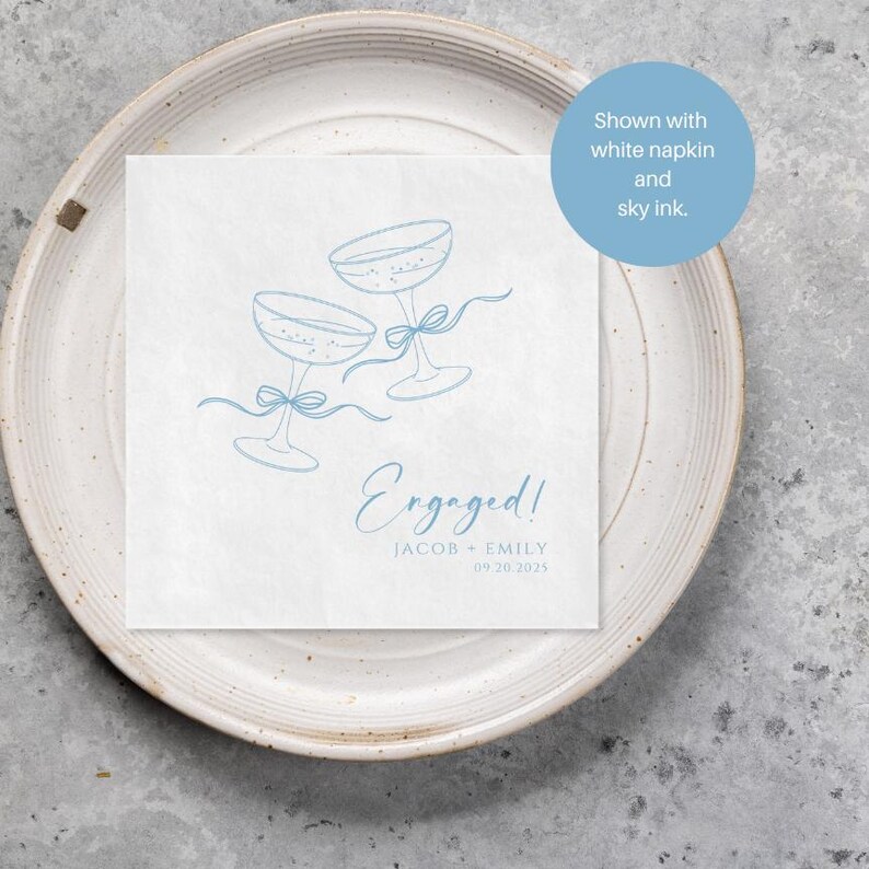 May include: A square white paper napkin featuring a light blue line drawing of two champagne glasses tied with ribbons. The text reads Engaged! JACOB + EMILY 09.20.2025 in blue ink. A blue circular overlay states Shown with white napkin and sky ink.