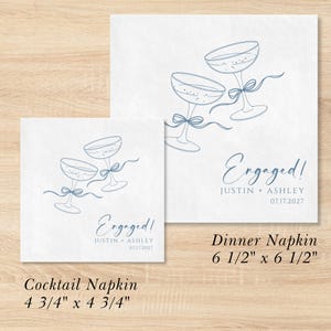 May include: Two white square paper napkins featuring blue line art of two champagne glasses tied with a ribbon. The text reads Engaged! Justin + Ashley 07.17.2027. The cocktail napkin measures 12.07 cm by 12.07 cm and the dinner napkin 16.51 cm by 16.51 cm.