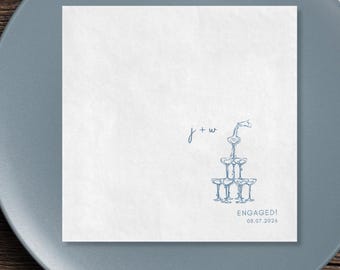 May include: White square paper napkin with a blue illustration of a champagne tower and the word "ENGAGED!" with a date. The napkin also features the initials "j + w" in a delicate script, perfect for a wedding or engagement celebration.