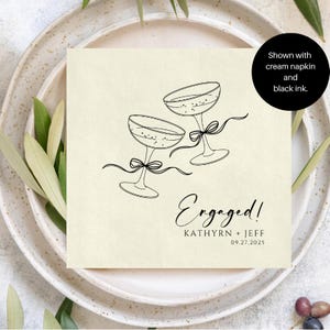 May include: A square cream-coloured napkin featuring a black line drawing of two champagne glasses tied with ribbons. The text reads "Engaged!", "KATHYRN + JEFF", and "09.27.2025". A black circular overlay states "Shown with cream napkin and black ink."