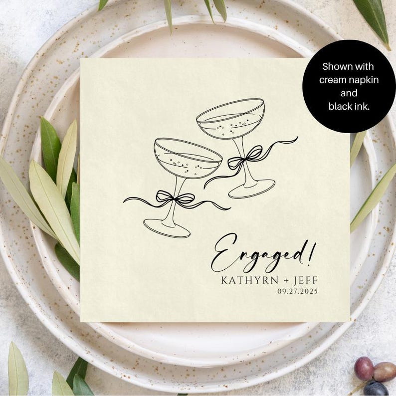 May include: A square cream-coloured napkin featuring a black line drawing of two champagne glasses tied with ribbons. The text reads "Engaged!", "KATHYRN + JEFF", and "09.27.2025". A black circular overlay states "Shown with cream napkin and black ink."