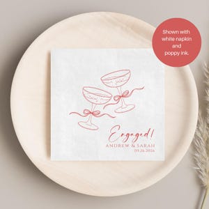 May include: A square white paper napkin featuring a red line art illustration of two champagne glasses tied with ribbons. The text reads Engaged! Andrew & Sarah 09.26.2026 in a red script and sans-serif font. The napkin is displayed on a round wooden plate.