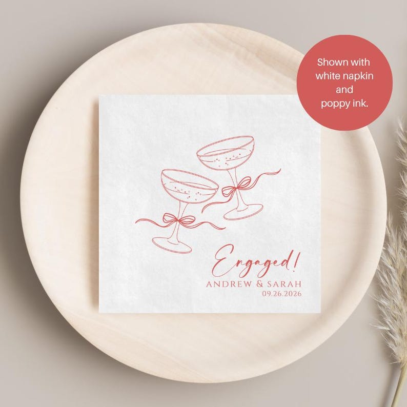 May include: A square white paper napkin featuring a red line art illustration of two champagne glasses tied with ribbons. The text reads Engaged! Andrew & Sarah 09.26.2026 in a red script and sans-serif font. The napkin is displayed on a round wooden plate.