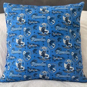 May include: A square blue pillow with a repeating pattern of Ravenclaw crests, banners, and text. The crests feature a raven, and the text reads "Ravenclaw". The pillow is likely made of fabric.