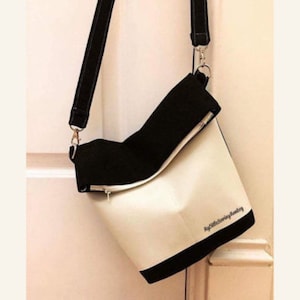 Puede incluir: Un bolso bandolera blanco y negro con una correa negra. El bolso tiene una solapa que se pliega y un cierre de cremallera. El bolso tiene el texto "High Society Handmade" impreso en la parte inferior.