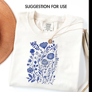 May include: Cream-colored t-shirt with a blue floral design. The shirt features a crew neck and short sleeves. A gold necklace with a small pendant rests on the shirt. The text "SUGGESTION FOR USE" is at the top.