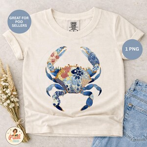 May include: A cream-colored short-sleeve t-shirt featuring a central graphic of a crab. The crab is decorated with various blue, white, and red floral and geometric patterns. The shirt has a crew neckline and a Comfort Colors brand label at the neck.