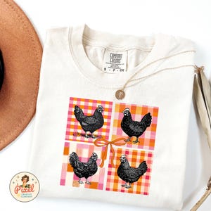 Puede incluir: Una camiseta de color crema con un gráfico de cuatro pollos negros en un patrón de cuadros rosa y naranja. El gráfico tiene un gran lazo naranja en el centro.