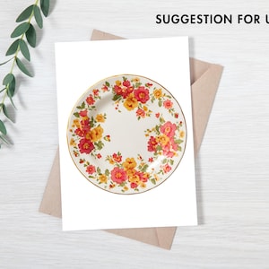 Pode incluir: Um prato de cer&acirc;mica branca com borda dourada, adornado com um padr&atilde;o floral em tons de vermelho, laranja e amarelo. O prato &eacute; apresentado em uma superf&iacute;cie branca com um envelope marrom e um galho de vegeta&ccedil;&atilde;o.